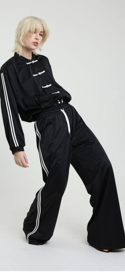 Pantaloni palazzo elastico da tuta neri con bande bianche, indossati da una modella con capelli biondi.